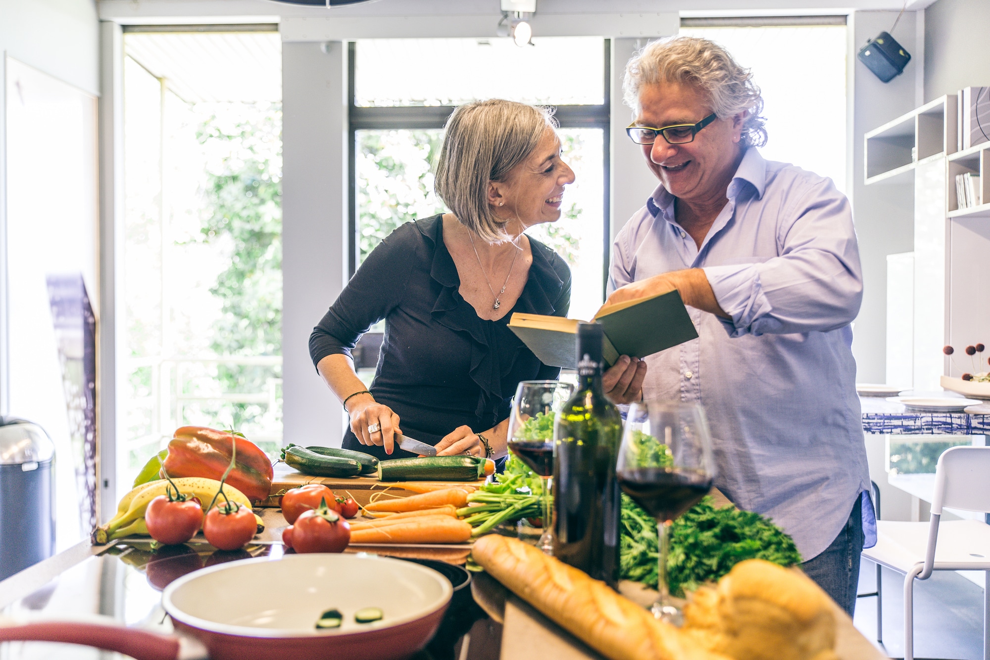 Senior couple cooking meal at home, man reading a cooking recipe book and woman cutting vegetables - Happy mature couple in the kitchen